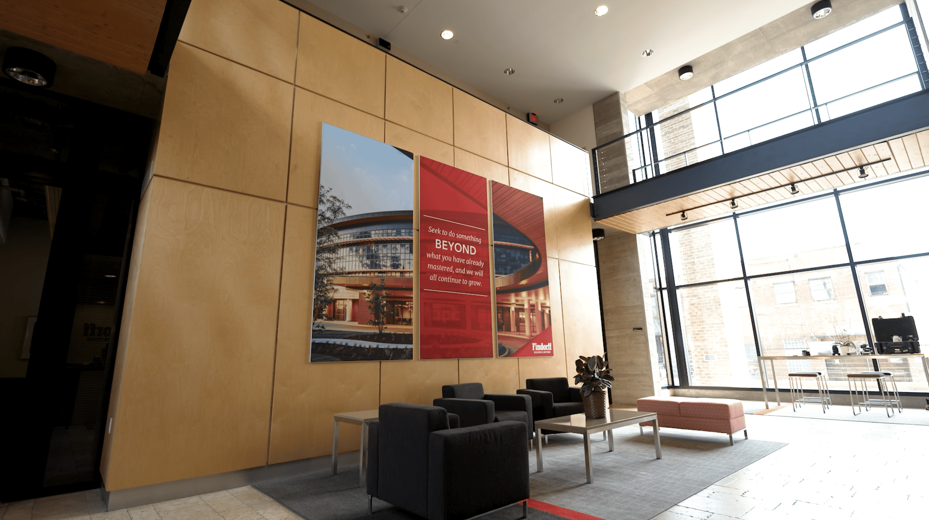 A modern lobby area with large wooden wall panels featuring a multi-panel graphic display. The left panel shows an architectural photo of a building, while the right panel has a red background with the text: "Seek to do something BEYOND what you have already mastered, and we will all continue to grow." Below, the Findorff logo reads "Building & Beyond."